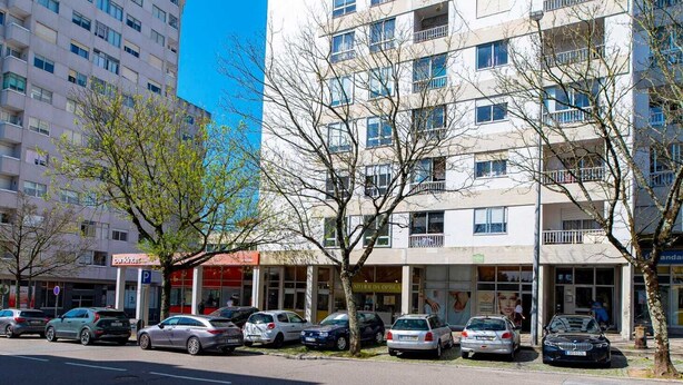 Um edifício de vários andares com varandas, carros estacionados e um céu azul límpido.