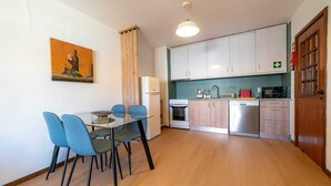 Uma cozinha compacta com armários brancos, micro-ondas, lava-louças e um espaço de refeições com mesa de vidro e cadeiras azuis.
