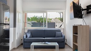 Una sala de estar moderna con un sofá gris, una mesa de centro blanca, un televisor montado en la pared y un ventanal con vistas a árboles y edificios.