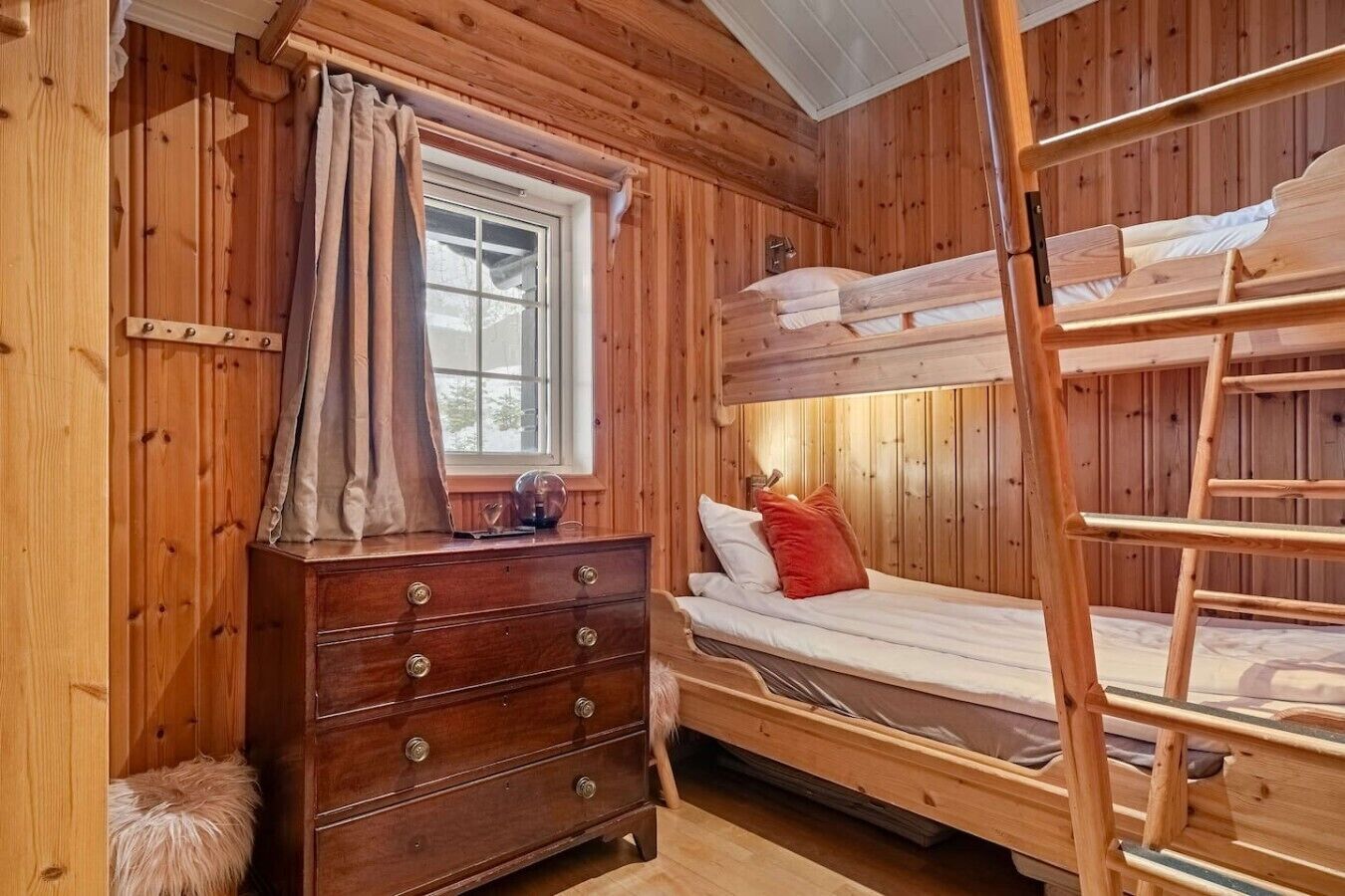 A wooden cabin room with a bunk bed, a dresser, and a window with curtains.