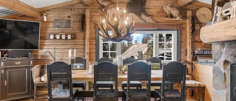 A rustic wooden interior with a dining area featuring a large table and chairs, a mounted bear head, and a stone fireplace.