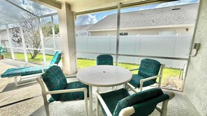 A covered patio with a round table and four chairs, a bench, and a view of a house and yard. - Sunlit Splash Private Pool (Kissimmee)