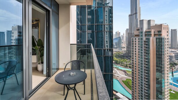 Un balcon doté d’une balustrade en verre, d’une table et de chaises, offrant une vue sur un paysage urbain avec de hauts immeubles et une piscine.