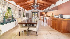 Uma sala de jantar espaçosa com uma longa mesa de madeira e cadeiras, uma cozinha com armários de madeira e um micro-ondas, e um teto com vigas aparentes.