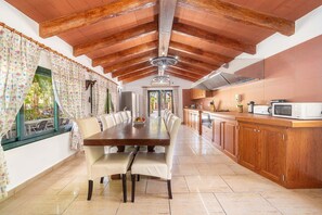 A spacious dining area with a long wooden table and chairs, a kitchen with wooden cabinets and a microwave, and a ceiling with exposed beams. - Can Tirenet accommodate 8 people Wi-Fi, swimming pool, tennis/padel (Alcudia)