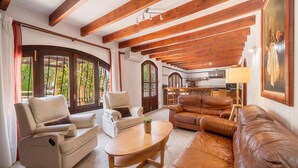 Sala de estar com sofá, poltronas, mesa de centro, vigas de madeira, cozinha ao fundo e grandes janelas.