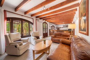 A living room with a sofa, armchairs, a coffee table, wooden beams, a kitchen in the background, and large windows. - Can Tirenet accommodate 8 people Wi-Fi, swimming pool, tennis/padel (Alcudia)