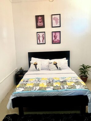Un dormitori amb llit, almoines amb motius florals, una nit del llit amb una planta i obra d'art mural amb figures africanes i un mapa.
