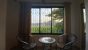 A balcony with two wicker chairs and a small round table, overlooking a green field. - GFV Stays (Candolim)