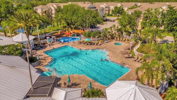 A large outdoor swimming pool surrounded by palm trees and lounge chairs, with a playground and a covered seating area nearby. - Universal Palms Retreat at Windsor Palms (Kissimmee)