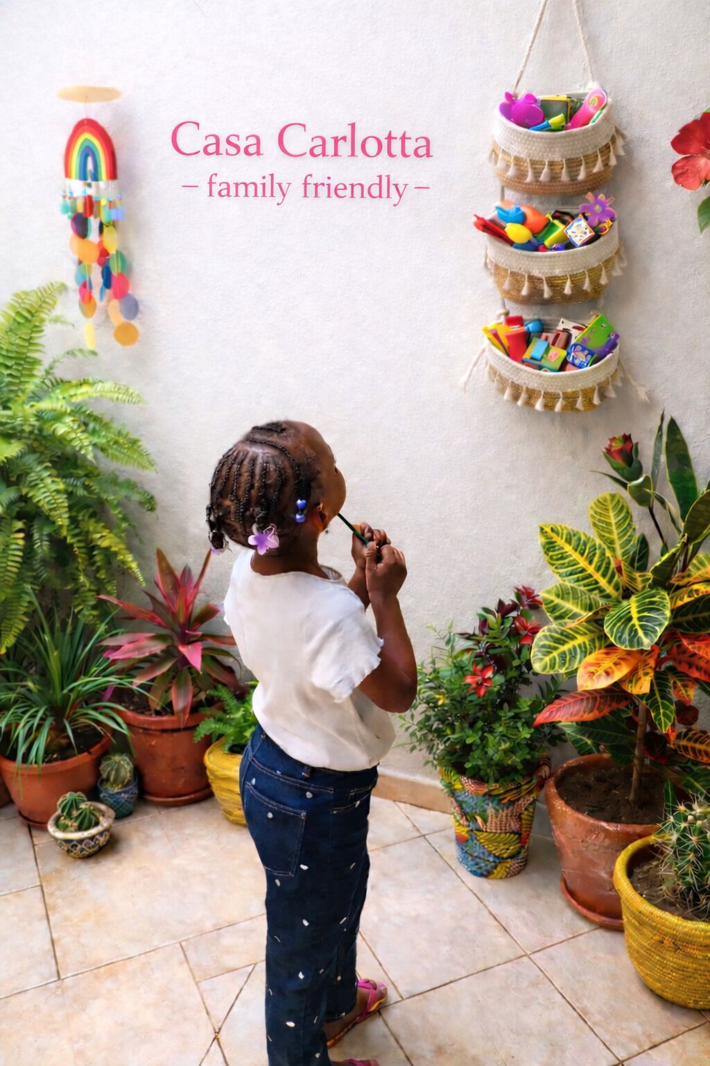 Un enfant vêtu d’une chemise blanche et d’un jean bleu, entouré de plantes en pot et d’un panier suspendu contenant des objets colorés.