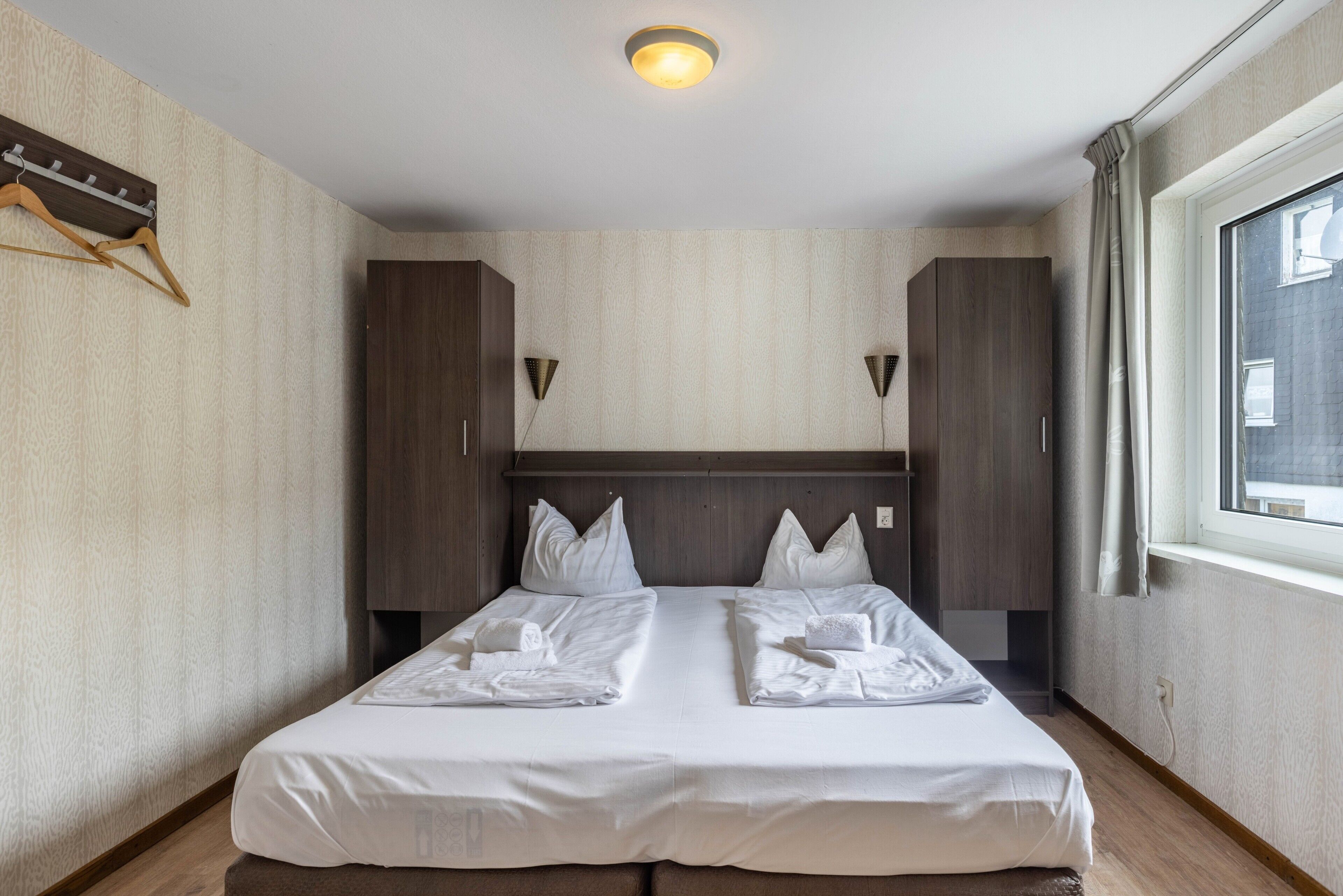 Ein Hotelzimmer mit einem Doppelbett, zwei Nachttischen, einem Kleiderschrank, einem Fenster mit Vorhängen und einer Wandleuchte.