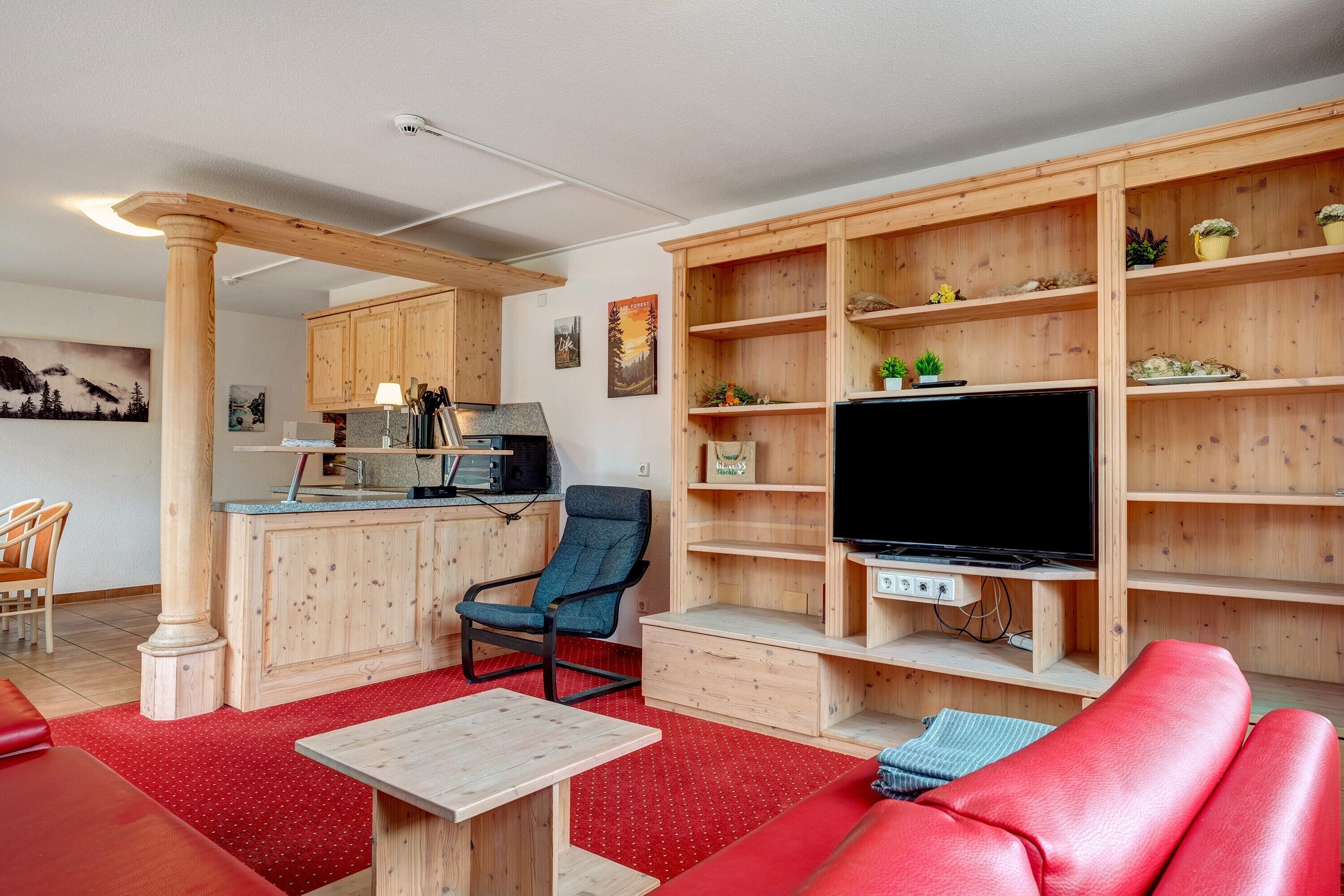 A living room with a red sofa, a TV, wooden shelves, a kitchen area, and a dining table.