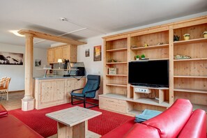 A living room with a red sofa, a TV, wooden shelves, a kitchen area, and a dining table.