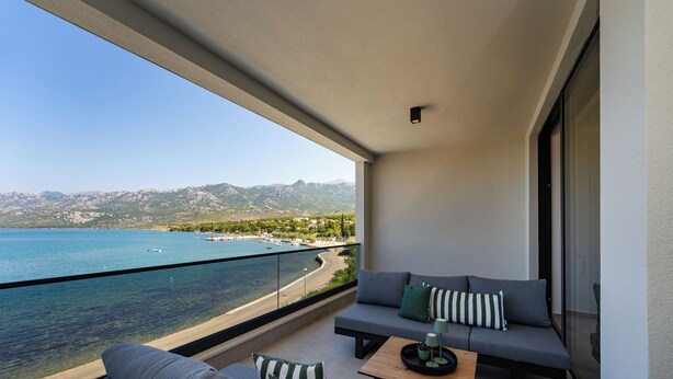 Ein Balkon mit einer Couch, einem Sessel, einem kleinen Tisch und Blick auf Strand und Berge.