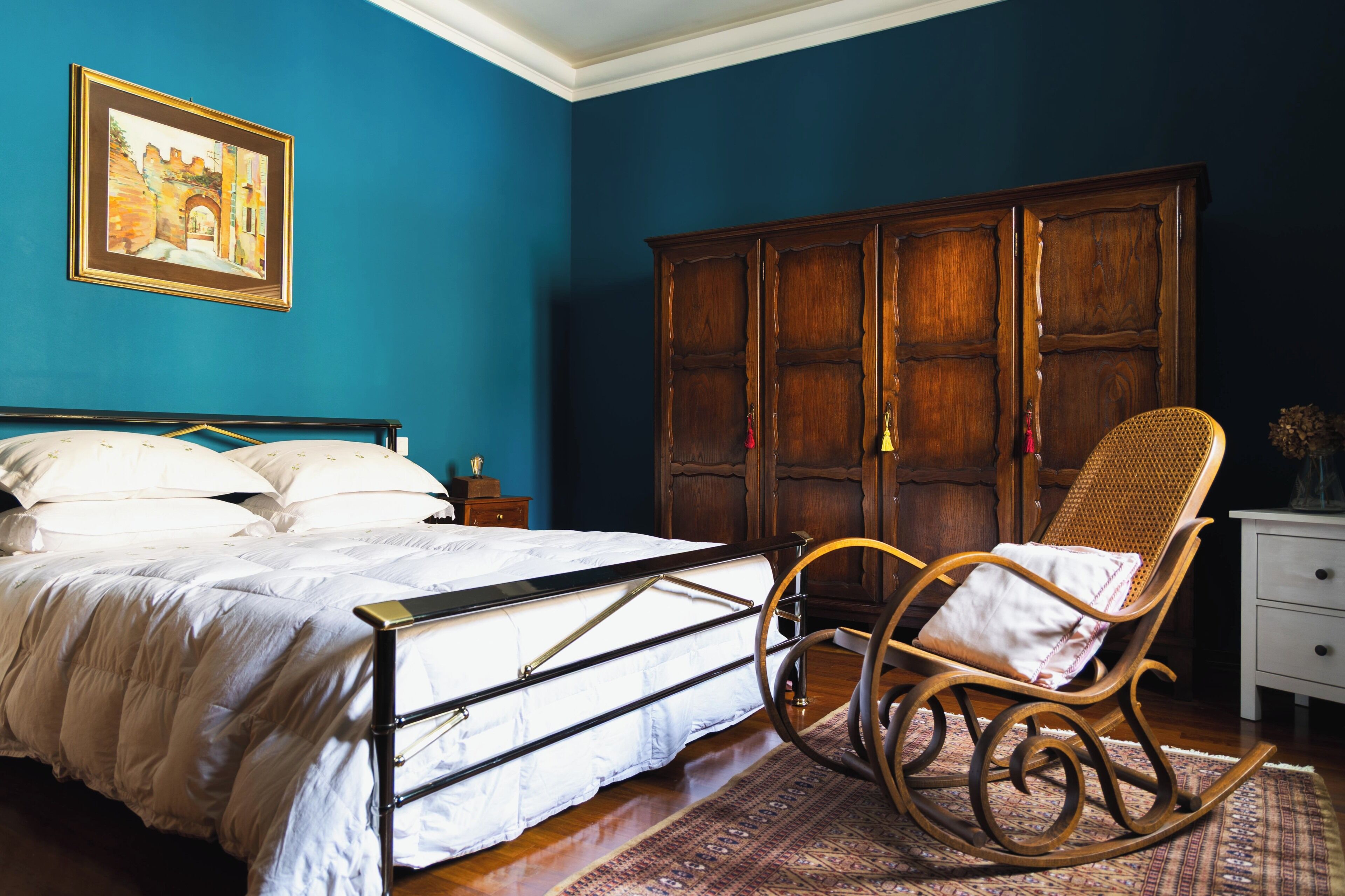 A bedroom with a bed, a rocking chair, a wardrobe, and a painting on the wall.