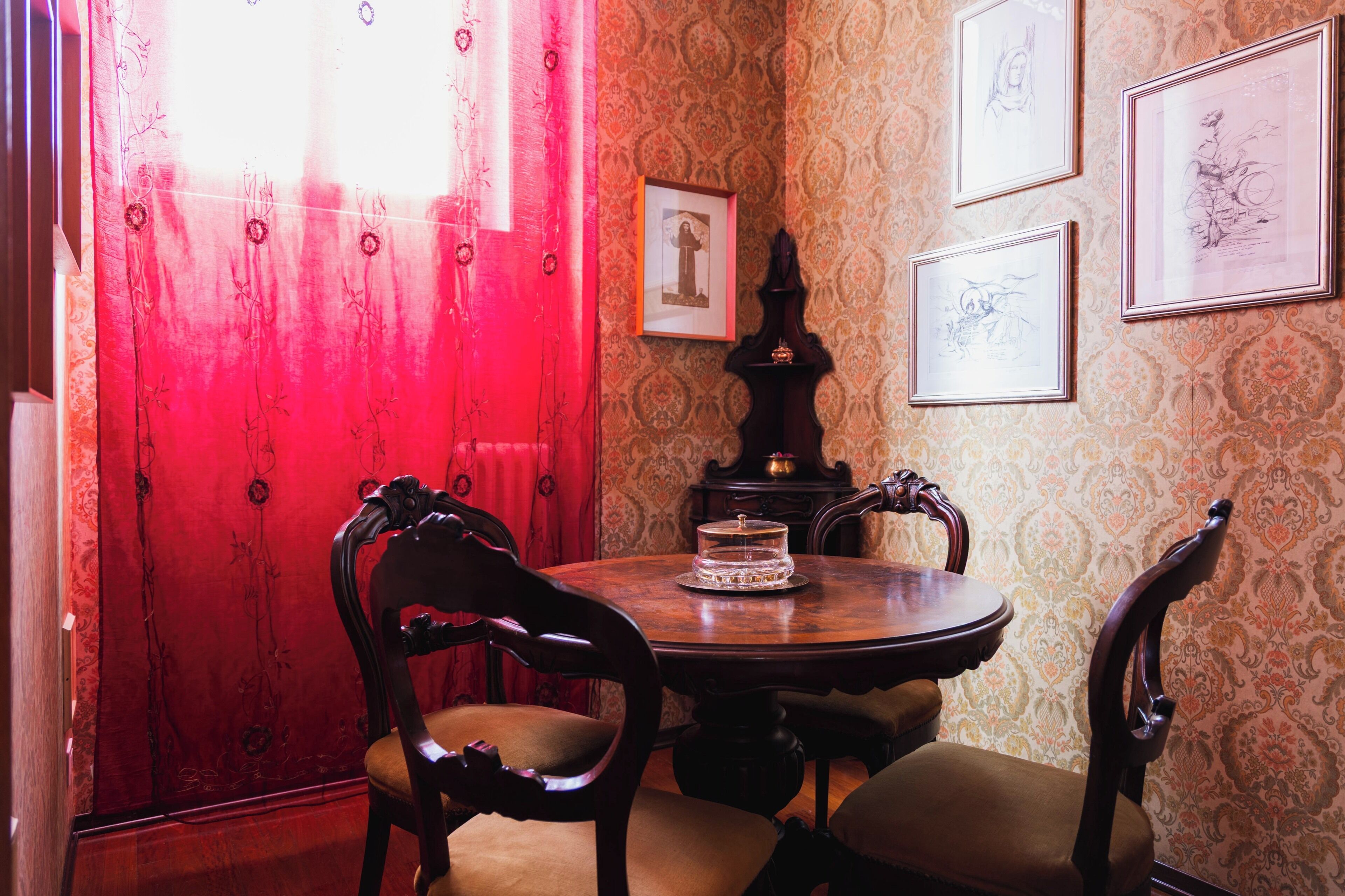 Un comedor con una mesa redonda, cuatro sillas, una cortina roja y tres obras de arte enmarcadas en la pared.
