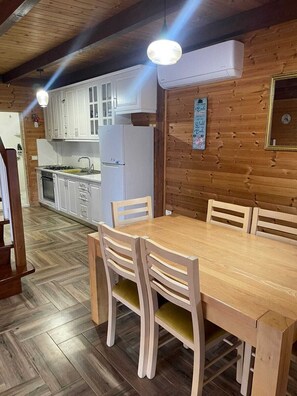 A wooden interior with a dining table and chairs, a kitchen area with white cabinets, and a wall-mounted clock.