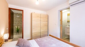 Ein Schlafzimmer mit Bett, Kleiderschrank und einem durch eine Glastür einsehbaren Badezimmer sowie einer kleinen Pflanze.
