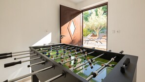 Ein Tischkicker in einem Raum mit einer Tür zu einem Außenbereich mit Pool und Liegestühlen.