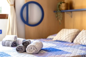 Uma cama com toalhas dobradas, um ornamento circular azul na parede e uma prateleira de madeira com uma planta.