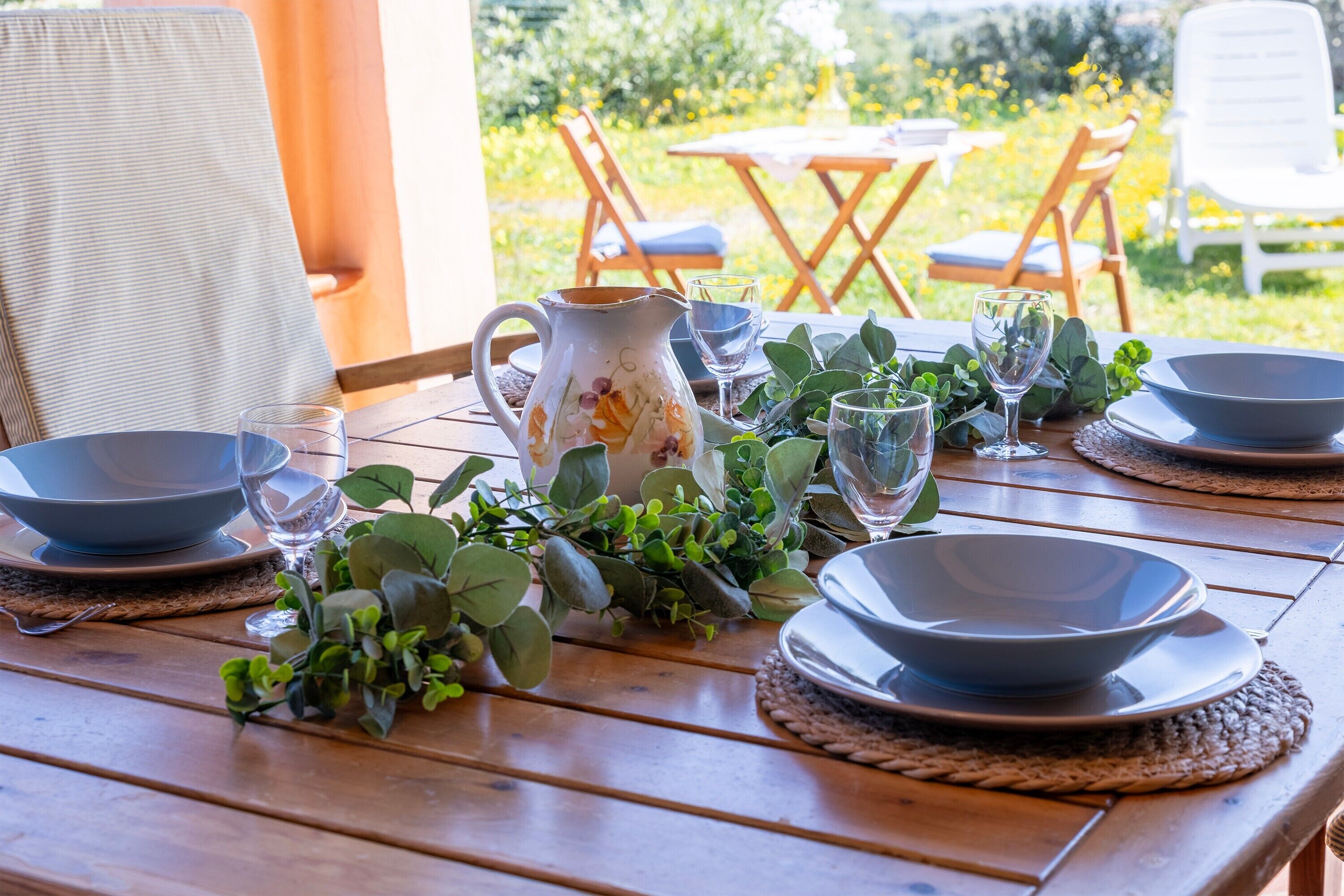 Une table en bois, dressée pour le repas, avec des bols, des verres et une carafe, le tout entouré de verdure.