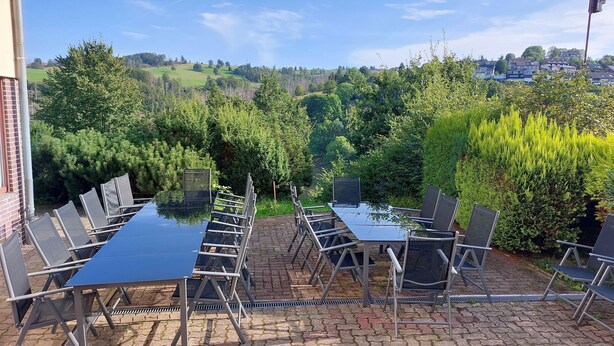 Eine Terrassensitzgelegenheit im Freien mit einem langen Tisch und Stühlen, umgeben von viel Grün und Blick auf eine Stadt.