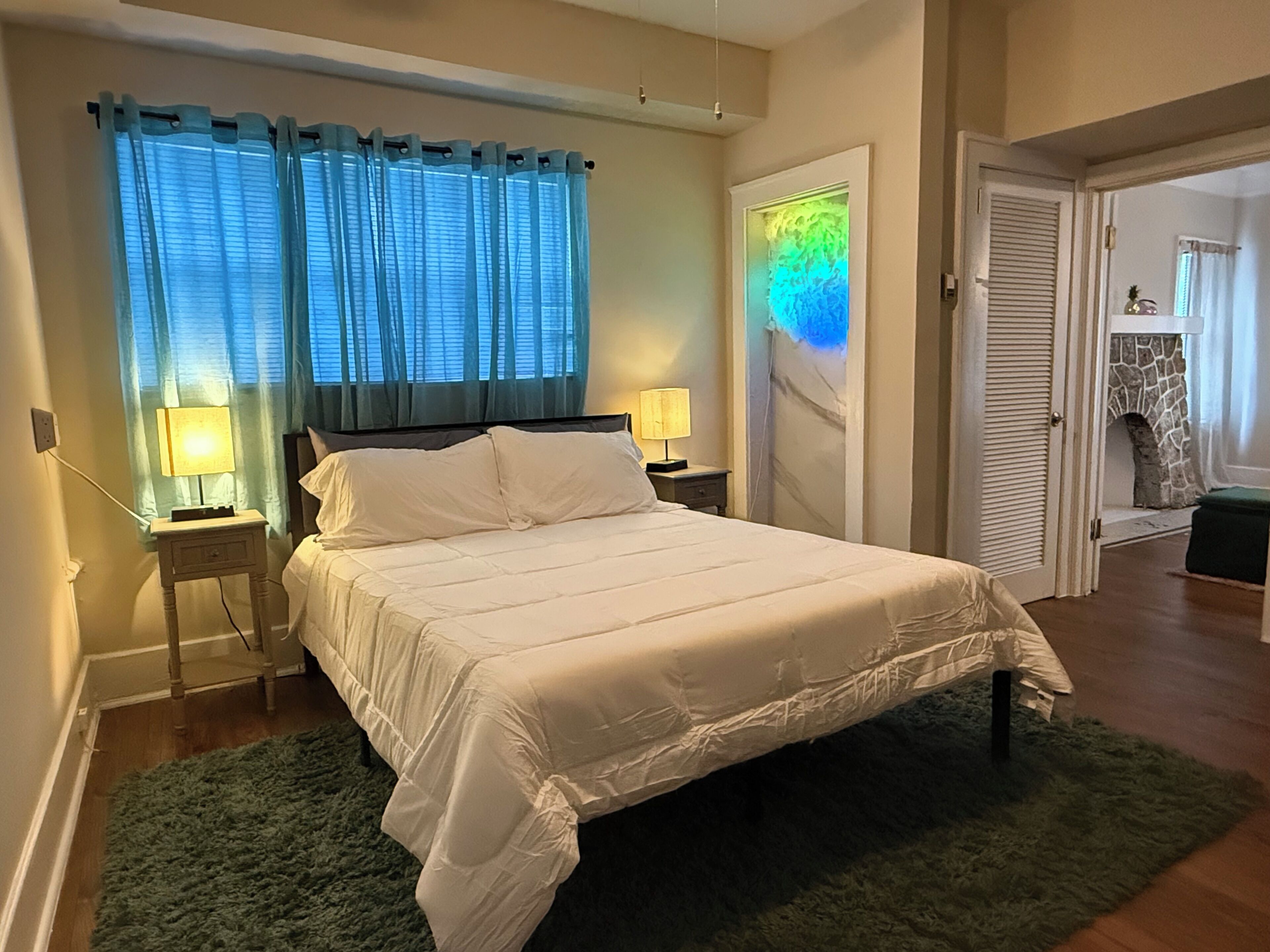 A bedroom with a bed, two bedside lamps, a window with curtains, a wall-mounted artwork, and a door leading to another room.