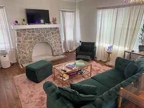 Un salon avec une cheminée en pierre, un canapé vert, une table basse avec des livres et un téléviseur posé sur une cheminée blanche.