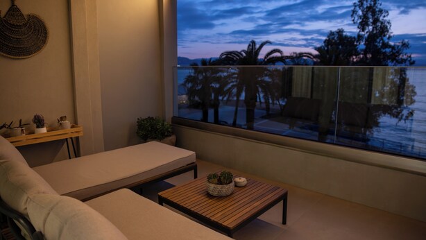 Ein Balkon mit Blick auf Palmen und einen Sonnenuntergang, eine Couch, ein kleiner Tisch mit einer Pflanze und ein dekoratives Wandelement.