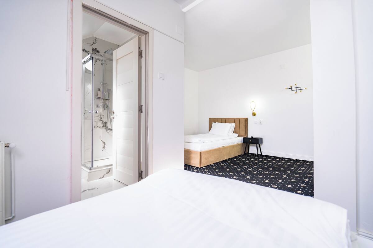 Habitación de hotel con cama, cabecero de madera, mesita de noche pequeña, luz de pared y zona de ducha.