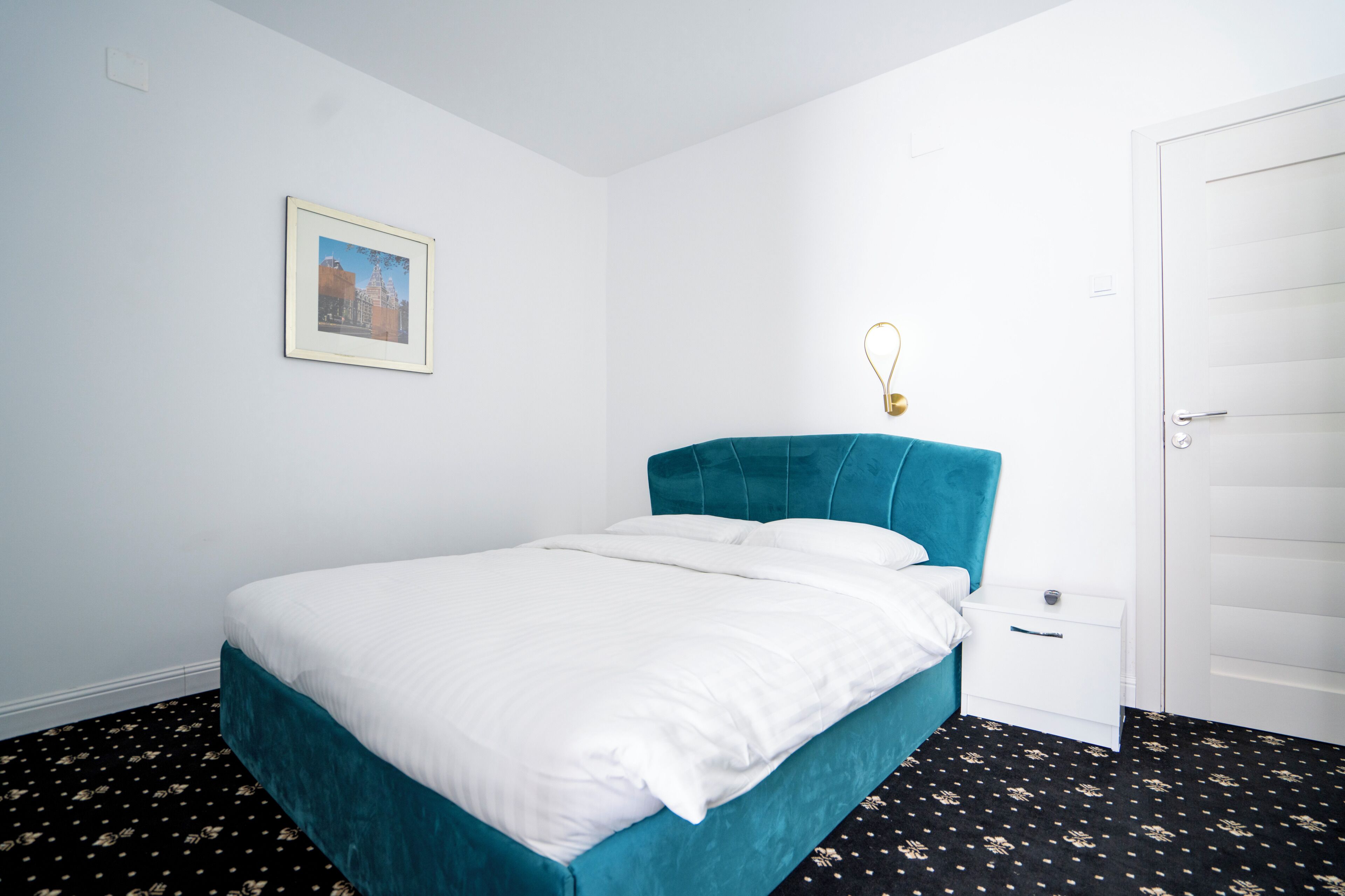 Una cama individual con cabecera color turquesa en una habitación con paredes blancas y piso con estampado blanco y negro.