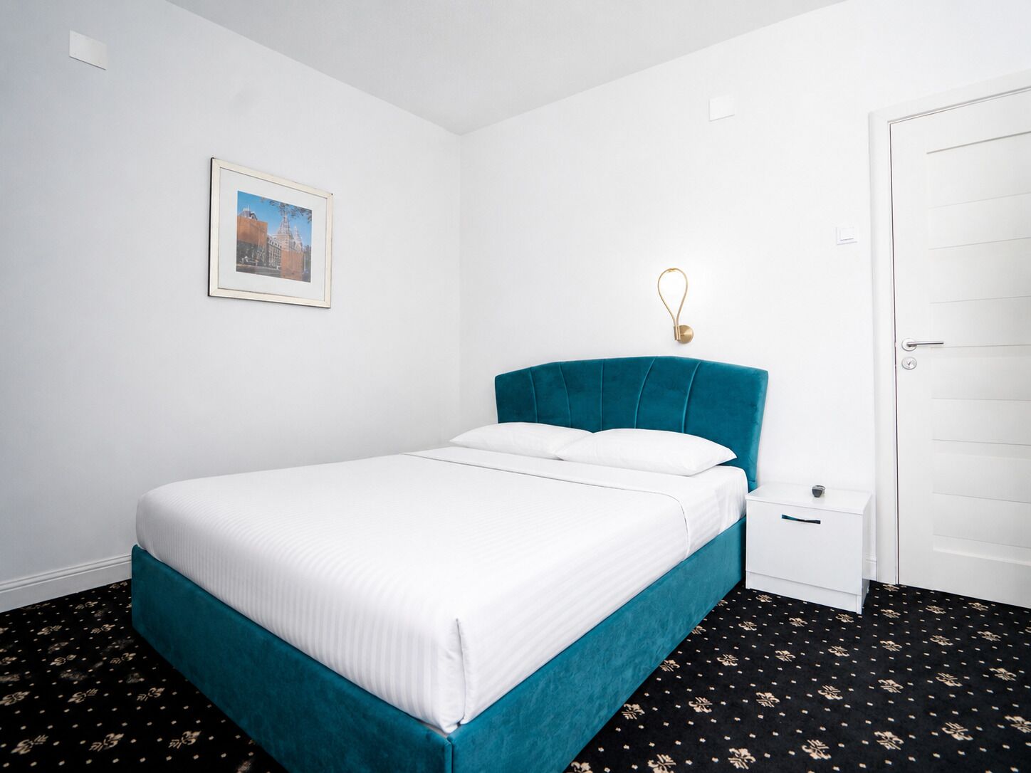 Un lit simple avec une tête de lit de couleur turquoise, une literie blanche, une petite table de chevet et une porte sur le côté droit.