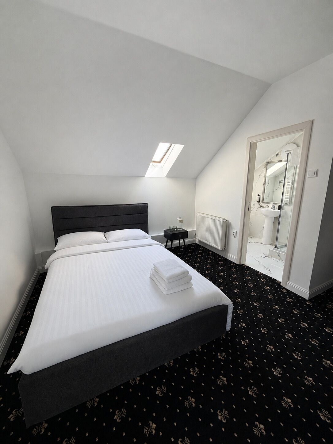 A hotel room with a bed, a small bedside table, a mirror, and a skylight.