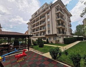 A multi-story residential building with balconies, a paved courtyard, and a small play area with a red horse-shaped toy. - Room-sunny Beach Central Apartment - 4 G (Nessebar)