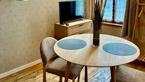 Ein Zimmer mit Fernseher auf einem Holztisch, einem runden Tisch mit zwei blauen Unterlagen, einem Stuhl und einem Fenster mit Vorhängen.