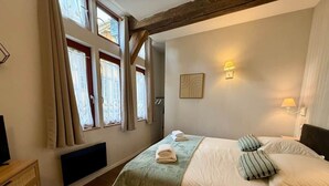 A bedroom with a bed, pillows, a bedside table, a window with curtains, and a wall-mounted lamp. - La Grande Escapade (T12) (Troyes)