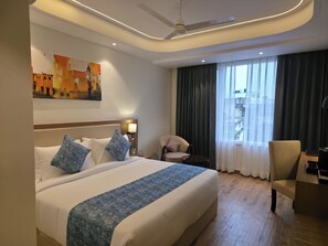 A hotel room with a bed, a chair, a desk, a painting, and a view of the city. - Lords Inn Katra (Katra)