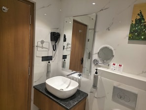 A modern bathroom with a marble wall, a wooden door, a white sink, a mirror, and a shower area. - Lords Inn Katra (Katra)