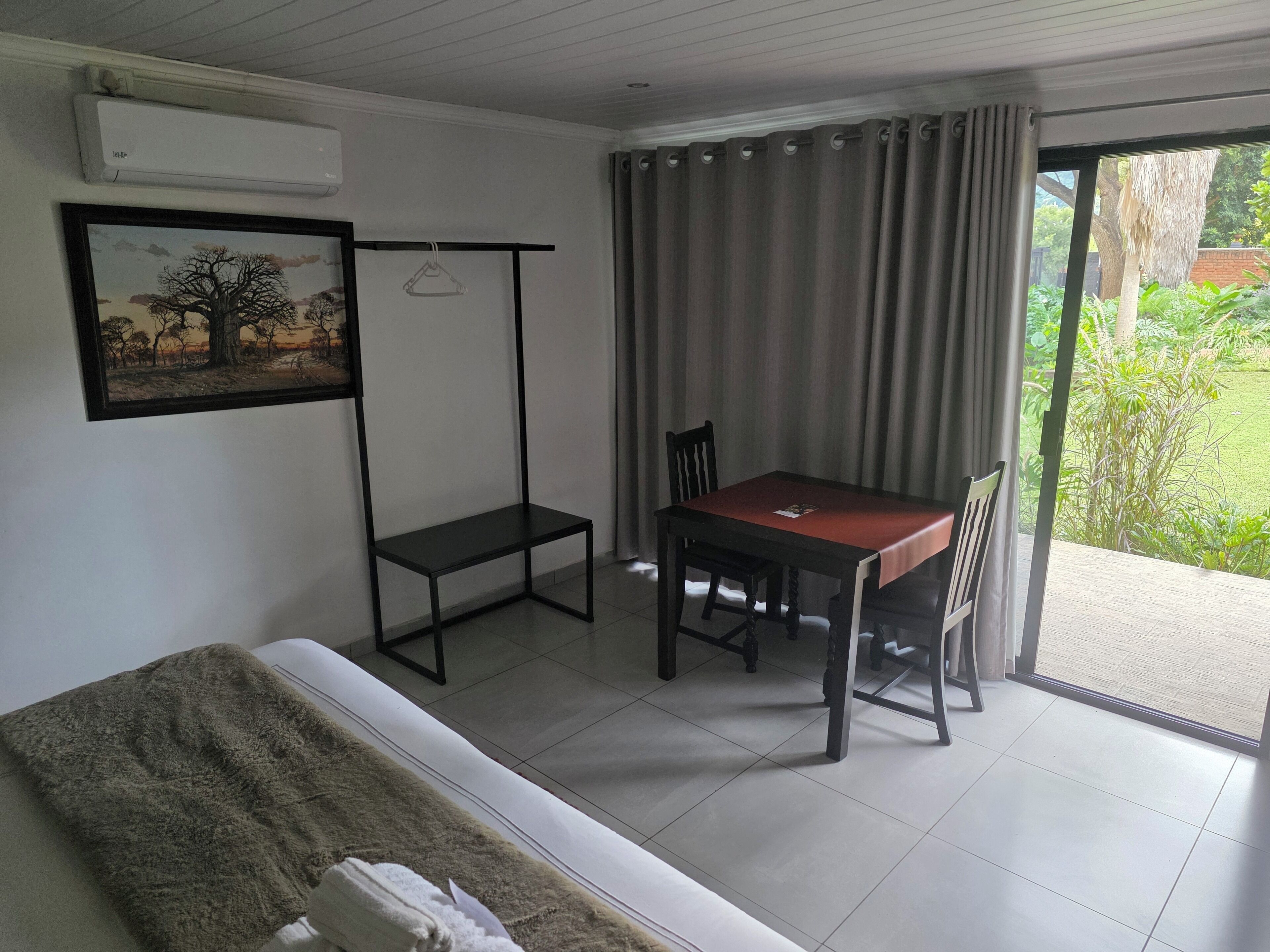 A room with a bed, a table with chairs, a hanging chair, a painting, and a view of greenery outside.