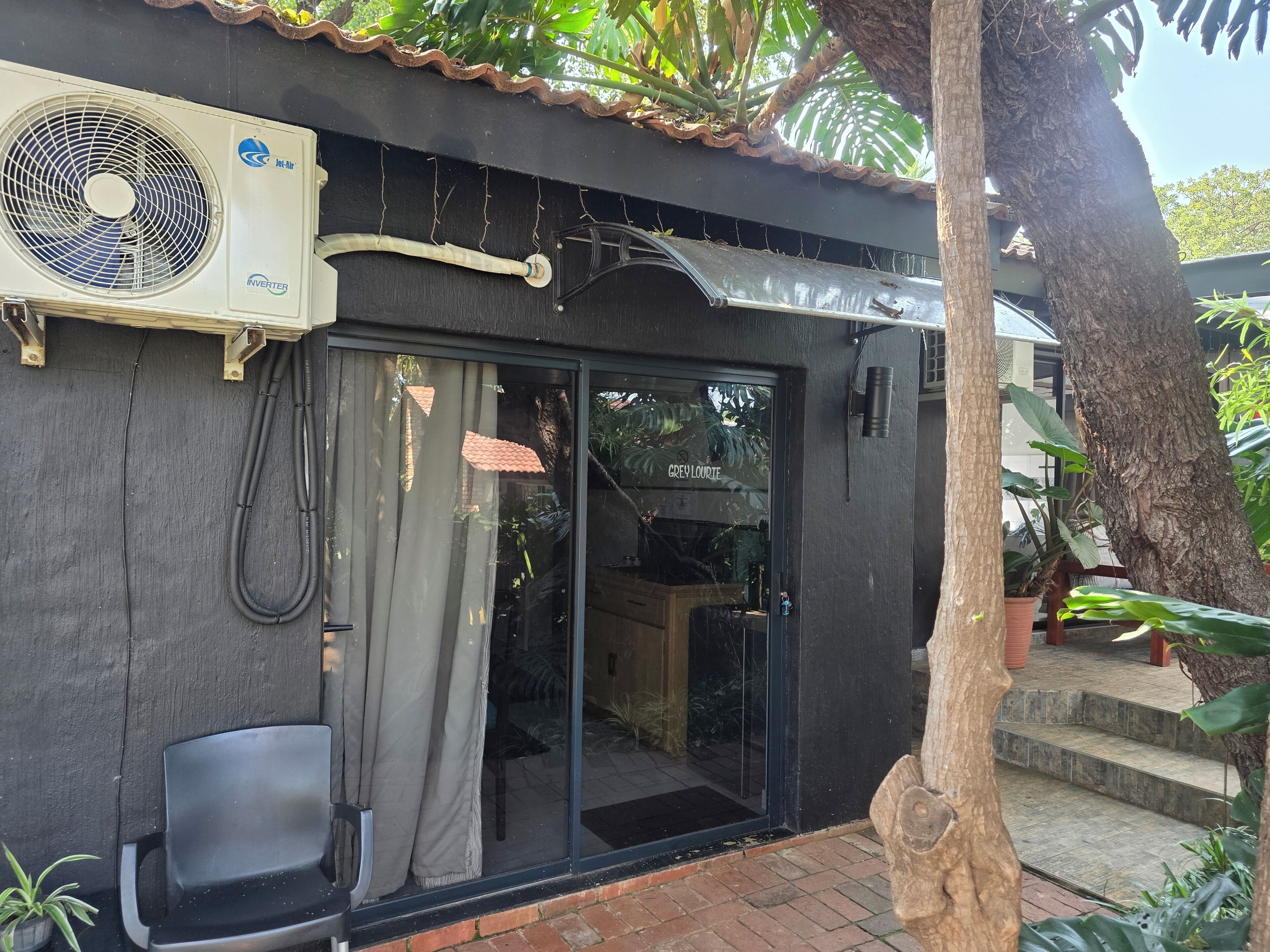 A building with a black exterior, a large air conditioning unit, a sliding glass door, and a small outdoor seating area with two chairs.