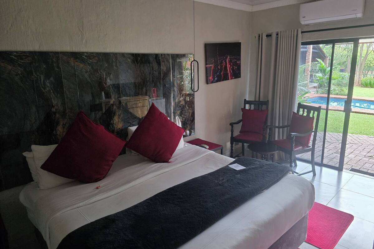 Habitación de hotel con una cama grande, almohadas rojas, cabecera de mármol, un cuadro, una silla y vista a una piscina.