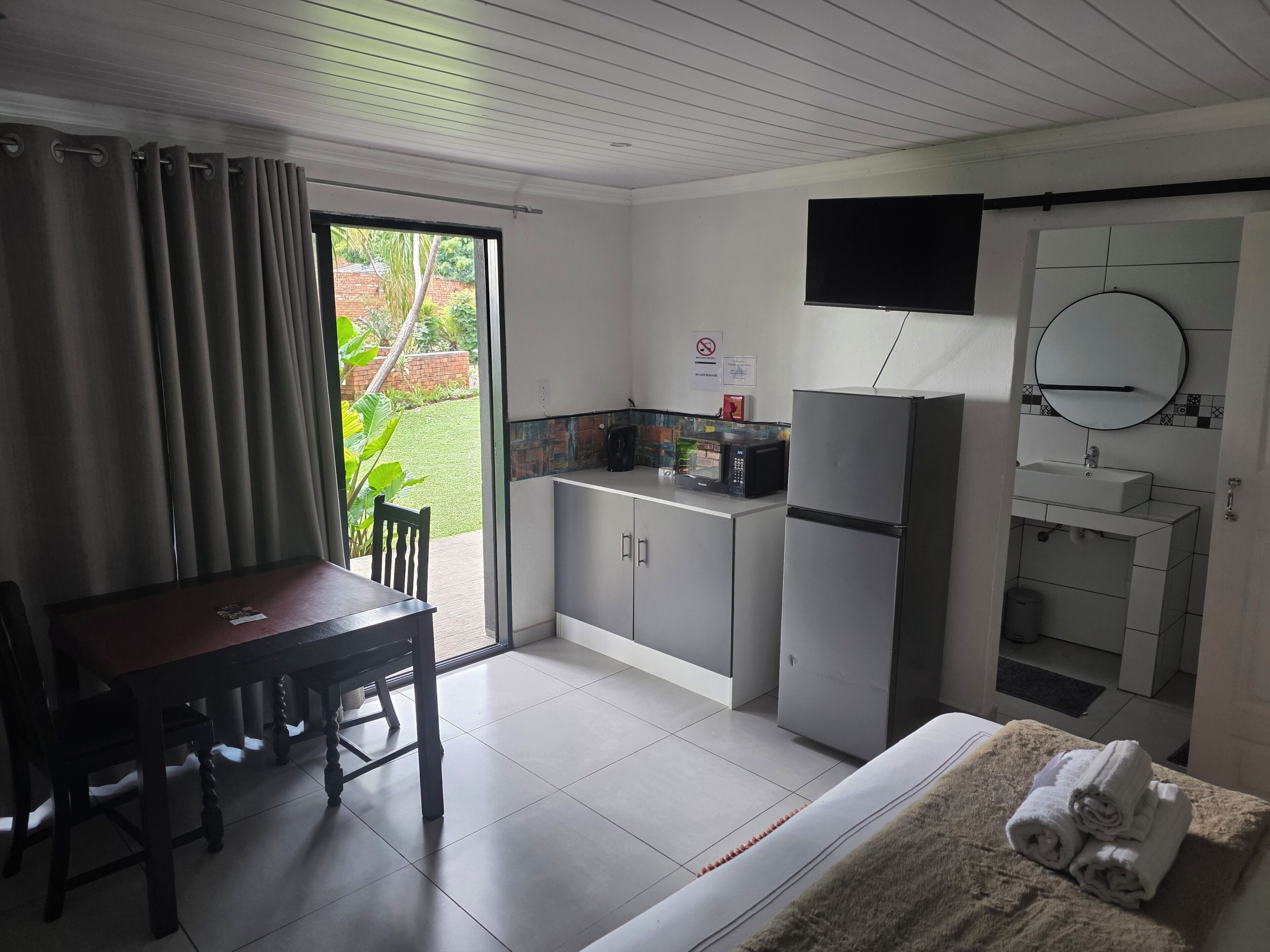 A compact hotel room with a dining table, a small kitchenette, a flat-screen TV, and a bathroom with a sink and mirror.
