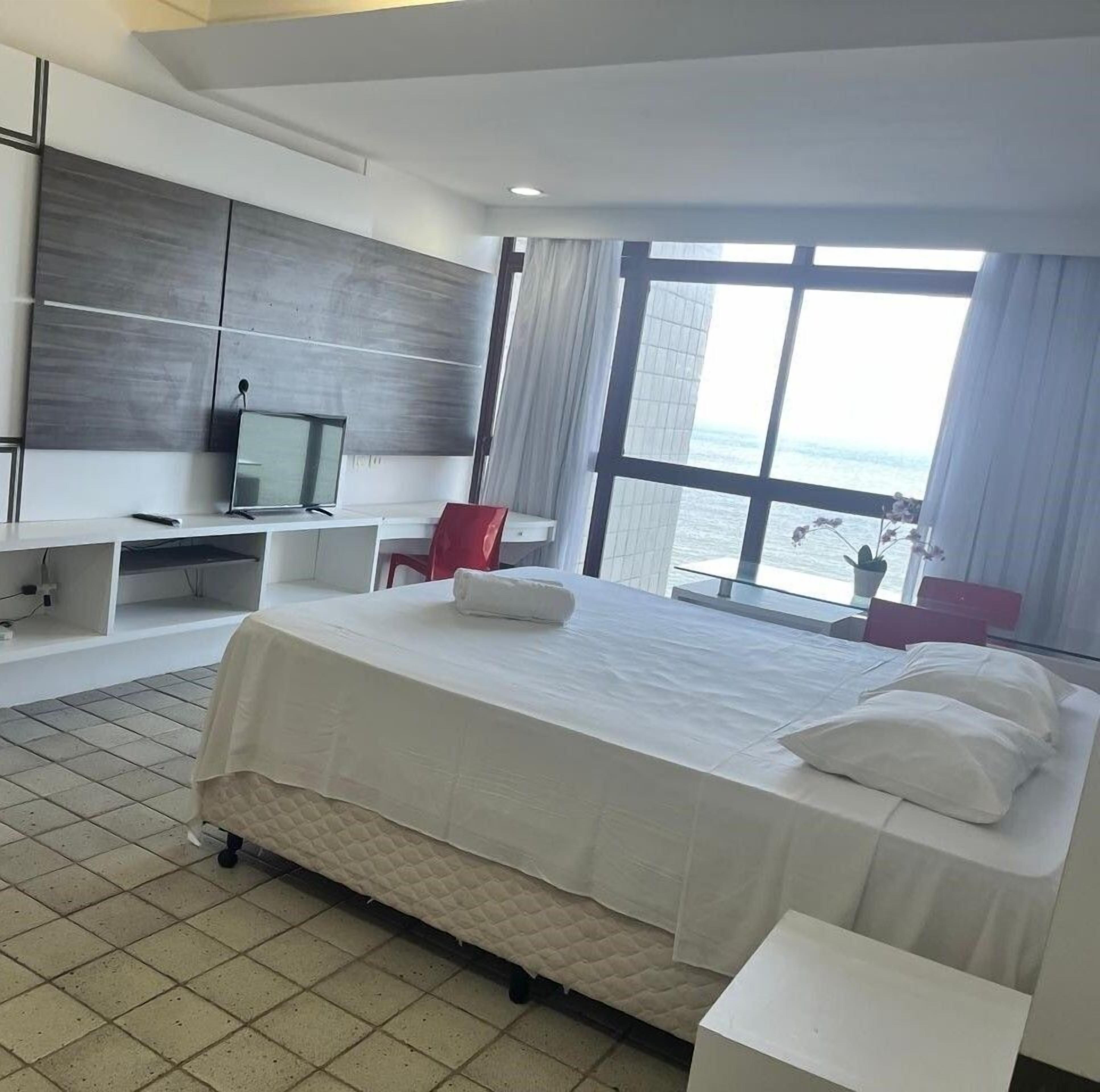 Quarto de hotel com cama, escrivaninha, televisão e uma grande janela.