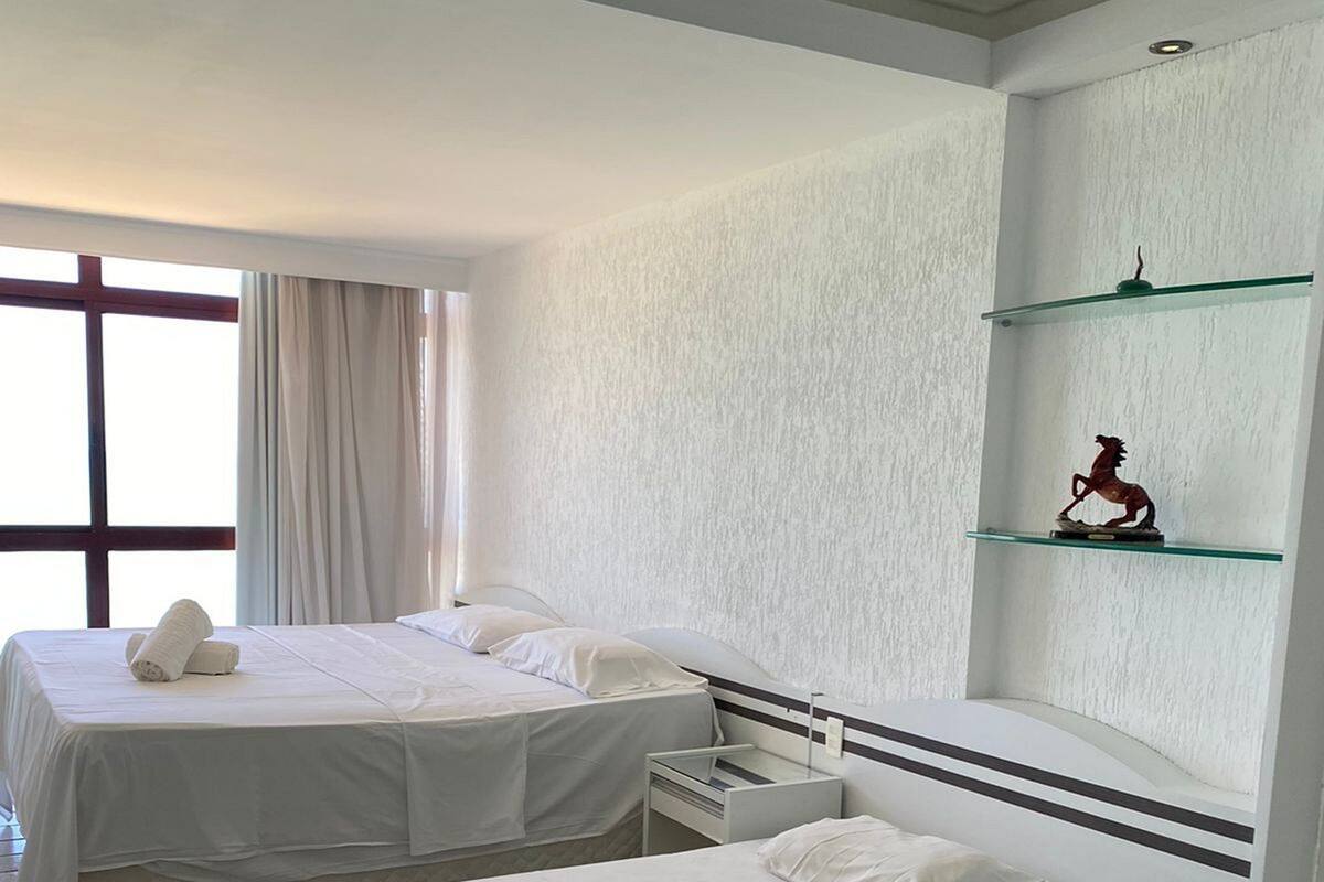 Habitación de hotel con dos camas, una mesita de noche pequeña, un estante en la pared con una estatua y un ventanal con cortinas.