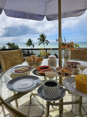 Una mesa dispuesta para el desayuno, con vista al océano, incluyendo platos, tazones y una cafetera.