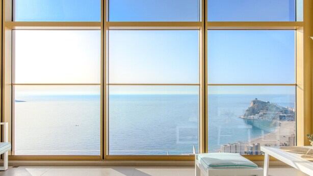 Ein Zimmer mit großen Fenstern und Meerblick, eine weiße Bank und ein kleiner Tisch mit einem Handtuch.