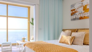 Ein Schlafzimmer mit großem Fenster, einem Bett mit weiß-orangefarbenem Bettbezug und Blick auf das Meer.