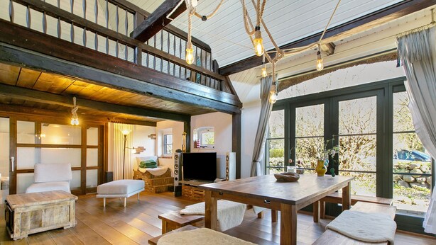Una habitación amplia con piso de madera, una gran mesa de madera, bancos, un sofá, un televisor y una escalera que sube a un nivel superior.