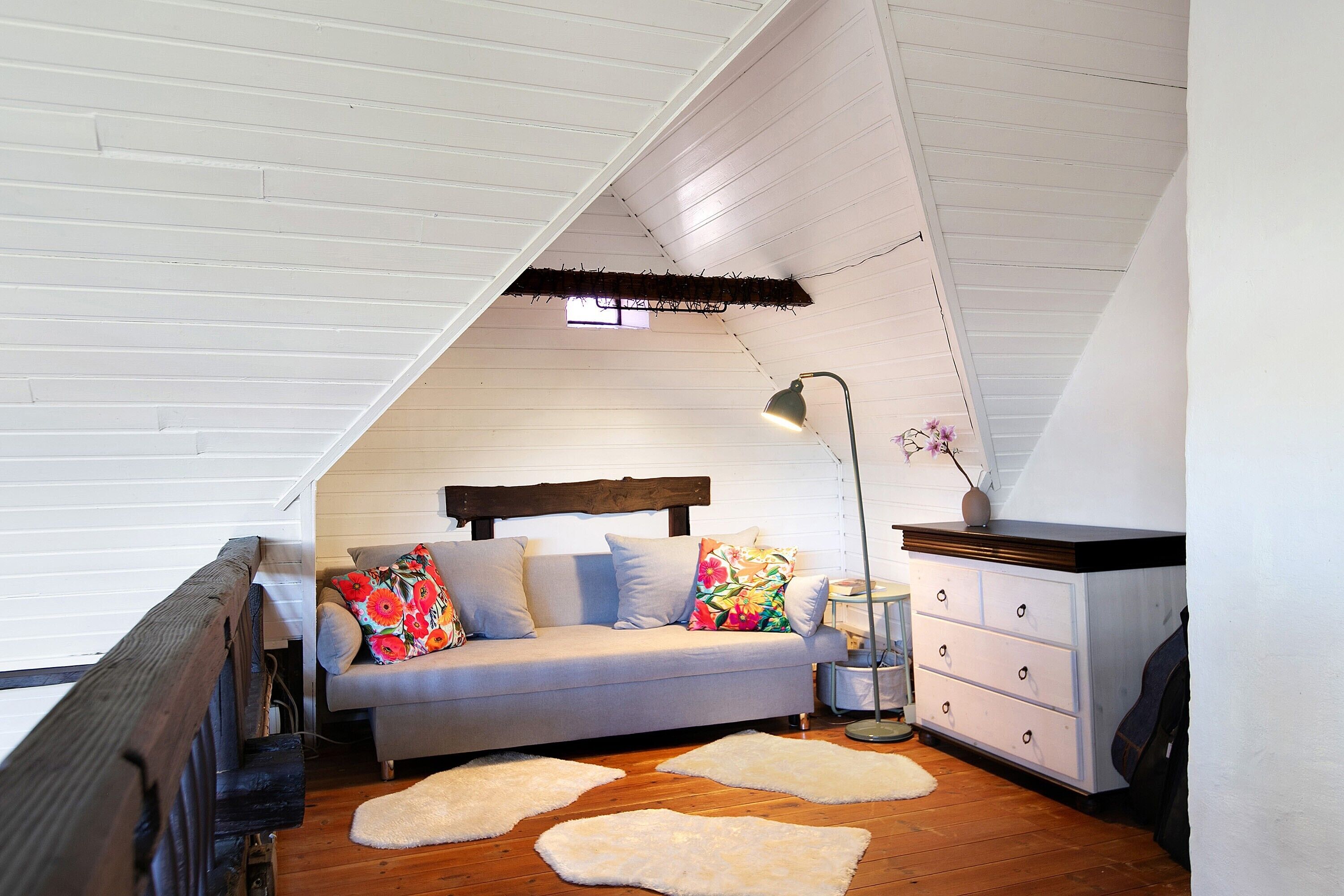 Ein Schlafzimmer im Loft-Stil mit schrägem Dach, einem Schlafsofa mit Blumenkissen, einem Schminktisch, einer Lampe und einem Fenster.